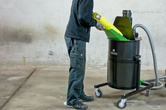 A person in a black jacket and cargo pants is pouring a self-levelling compound into a black mixing station with wheels, standing on a concrete floor.